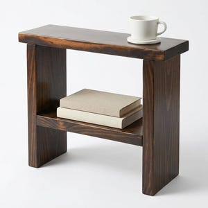 May include: A dark brown wooden side table with a lower shelf holding two books. A white mug and saucer sit on top. The table is stained and has a simple, modern design.