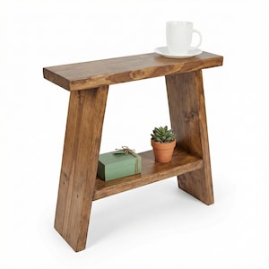 May include: A rustic, wooden side table with a lower shelf. A small potted succulent and a green box with a bow sit on the shelf. A white teacup and saucer rest on the table's top surface. The table has a warm, brown finish.