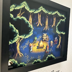 May include: A framed pixel art diorama depicting a campfire scene in a forest. The artwork features characters gathered around a fire, with trees and foliage in the background. The frame is black, and the artwork is signed by "FANBIT CREATIONS".