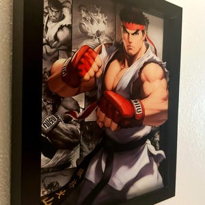 May include: Framed artwork featuring a dynamic illustration of a martial arts character in a white gi and red gloves, with a red headband. The artwork is set against a black and white collage background. The frame is black.