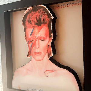 May include: Framed artwork depicting David Bowie from the Aladdin Sane album cover. The image shows Bowie with red hair, a lightning bolt across his face, and the album title. The artwork is in a black frame.
