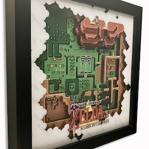 May include: A framed 3D shadow box display featuring a pixel art map from The Legend of Zelda: A Link to the Past. The artwork includes a green landscape, brown mountainous areas, and the game title logo with a sword graphic in a black frame.