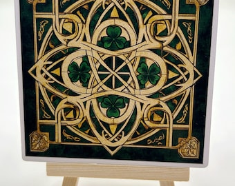 May include: A square ceramic tile with a green background and a gold Celtic knot design featuring four shamrocks. The tile is on a small wooden easel.