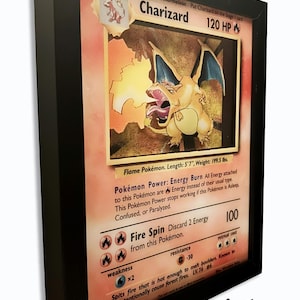 Peut inclure: Un cadre en relief 3D présentant une carte Pokémon Dracaufeu. La carte illustre la créature orange de type dragon crachant du feu, avec des textes incluant 120 PV, Danse Flamme et Pokémon Flamme. Le cadre noir met en valeur l'illustration en couches.
