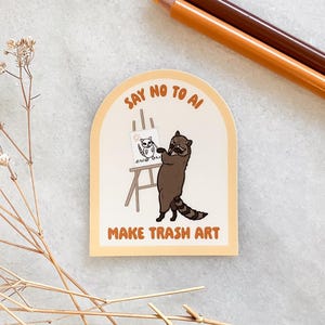 May include: A sticker featuring a cartoon raccoon painting on an easel. The sticker reads "SAY NO TO AI" and "MAKE TRASH ART". The raccoon is brown with a striped tail. Two pencils are in the background.