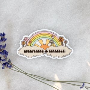 May include: A sticker with a rainbow design, sun, and flowers. The text "EVERYTHING IS TERRIBLE!" is printed on a cloud-shaped border. Dried lavender sprigs are in the image.