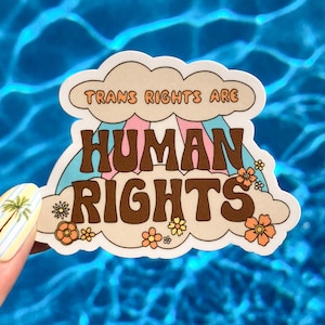 Peut inclure: Un autocollant avec le texte "TRANS RIGHTS ARE HUMAN RIGHTS" en lettres marron, rose et orange. Le design comprend un arc-en-ciel et des fleurs, sur un fond de nuage crème. L'autocollant est tenu sur un fond d'eau bleue.