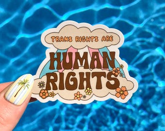 May include: A sticker with the text "TRANS RIGHTS ARE HUMAN RIGHTS" in brown, pink, and orange lettering. The design includes a rainbow and flowers, set against a cream-colored cloud background. The sticker is held against a blue water background.