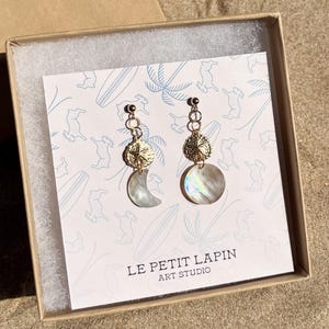 May include: Gold-coloured earrings with a moon and circular iridescent shell design. The earrings are displayed in a box with a card featuring a bunny and surfboard pattern. The card also has the text "LE PETIT LAPIN ART STUDIO."