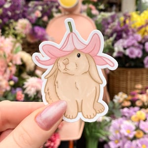 Peut inclure: Un autocollant représentant un lapin brun portant un chapeau de fleur rose. Le lapin est assis droit, les oreilles dressées. L'autocollant est sur un fond blanc.