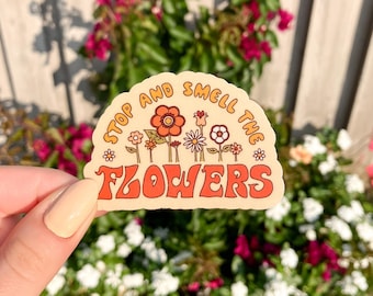 Peut inclure: Autocollant beige avec le texte "STOP AND SMELL THE FLOWERS" en orange et jaune. L'autocollant présente une rangée de fleurs stylisées en orange, blanc et jaune. L'autocollant a une esthétique rétro et groovy.