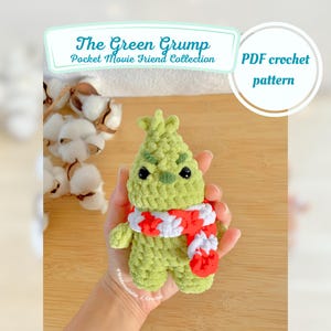 May include: A green crocheted Grinch amigurumi doll wearing a red and white striped scarf. The doll is being held in a hand. The text "The Green Grump Pocket Movie Friend Collection PDF crochet pattern" is visible in the image.