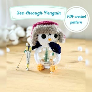 Op de afbeelding: Een gehaakte pinguïn met een doorzichtig lichaam, met een roze en grijze hoed en een marineblauwe sjaal. De pinguïn houdt miniatuurski's en skistokken vast. De tekst "See-through Penguin" en "PDF crochet pattern" worden weergegeven.