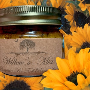 Op de afbeelding: Een pot Willow's Mist Pumpkin Pie Fall Body Scrub met een gouden deksel en een bruin etiket met een boomillustratie. De pot staat tegen een achtergrond van heldergele zonnebloemen.