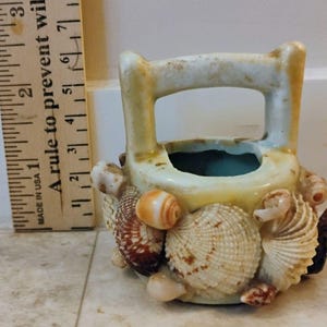 May include: A small, cream-colored ceramic basket-shaped vase, adorned with various seashells. The vase has a handle and a turquoise interior. The seashells are in shades of brown, white, and orange. The vase is approximately 3 inches tall.