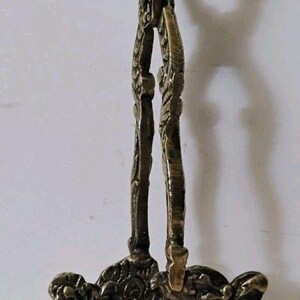 May include: A brass fireplace tool featuring a long handle with a decorative loop at the top and a wide, ornate base. The base is cast in a detailed relief pattern depicting a bearded face surrounded by intricate scrollwork and foliage motifs.