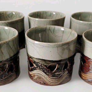 Op de afbeelding: Zes keramische bekers met een uniek ontwerp. De bovenste helft van elke beker is lichtgrijs met een craquelé-patroon, terwijl de onderste helft donkerbruin is met een gegraveerd golfpatroon. De bekers zijn in een cirkel gerangschikt.