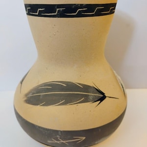 May include: A tan ceramic vase with a narrow neck and rounded base. The vase features a black geometric band around the rim, a painted gray feather motif on the center, and a solid black band near the bottom with a thin white arrow design.