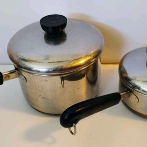 May include: Two stainless steel saucepans of different sizes, each featuring a polished silver finish, a black heat-resistant handle, and a matching lid with a black knob. The larger saucepan has a long handle, while the smaller one has a handle with a metal loop at the end for hanging storage.