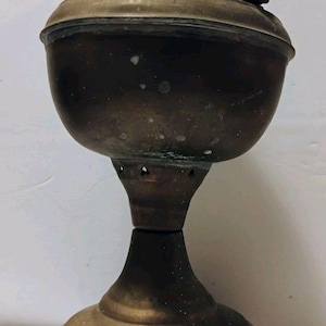 May include: An antique brass oil lamp featuring a wide, rounded reservoir, a flared pedestal base with small cross-shaped cutouts, and a top burner assembly with a horizontal adjustment knob. The metal surface shows signs of age and patina.