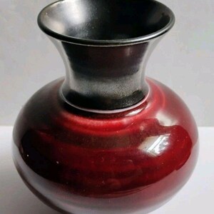 May include: A ceramic vase with a bulbous body and a flared neck. The main body features a deep, glossy red glaze with subtle horizontal banding. The neck and the interior rim are finished in a contrasting smooth, solid matte black glaze.