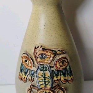 May include: A light green ceramic vase with a flared rim and a speckled finish. The front features a raised, relief-style design of a stylized bird or owl figure in shades of brown, yellow, blue, and green with intricate, carved-like details.