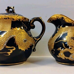 Könnte beinhalten: Eine schwarze Zuckerdose aus Keramik mit Deckel und ein passendes Milchkännchen. Beide Teile sind mit verzierten goldenen Drachenmotiven, goldenen Verzierungen an den Griffen und Rändern sowie goldenen geometrischen Symbolen versehen.