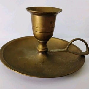 Peut inclure: Un bougeoir en laiton vintage avec une base ronde et une poignée. Le bougeoir a un sommet en forme de coupe pour tenir une bougie. Le laiton présente une patine chaude et vieillie, ajoutant à son charme antique. Une pièce classique pour la décoration intérieure.
