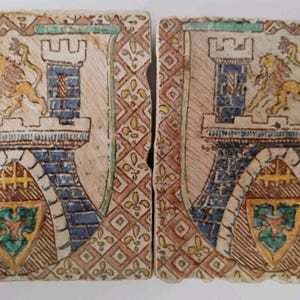 Puede incluir: Dos azulejos de cerámica cuadrados con un diseño heráldico pintado a mano de un castillo de piedra azul con dos torres y un león central. El borde muestra un patrón de diamantes repetitivo en rojo y beige con pequeños detalles de flores de lis.