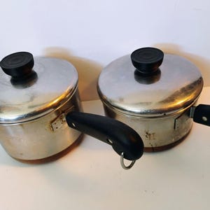 Puede incluir: Dos cacerolas de acero inoxidable con bases de color cobre, cada una con una tapa de acero inoxidable a juego con un pomo negro. Ambas cacerolas tienen mangos negros, uno de los cuales incluye una pequeña anilla metálica al final.