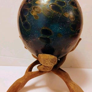 Op de afbeelding: Een bolvormig object met een donkerblauwe, groene en paarse gevlekte afwerking met ingebedde lichtgekleurde cirkelvormige patronen. De bol rust stevig in een gesneden houten standaard bestaande uit drie in elkaar grijpende, takachtige poten.