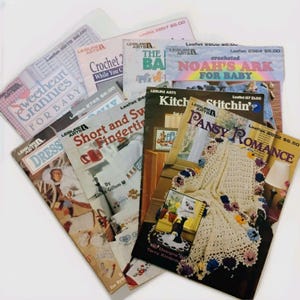 May include: A collection of vintage crochet and knitting pattern leaflets from Leisure Arts. Titles include "Sweetheart Grannies for Baby", "Crochet? While You Can", and "Pansy Romance". The leaflets feature colorful designs and illustrations.