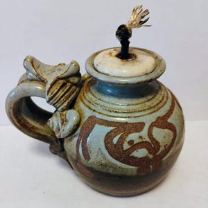 May include: Handmade ceramic oil lamp with a handle and lid. The lamp features a light blue and brown color scheme with decorative patterns. The wick is visible at the top. The lamp has a unique, artistic design.