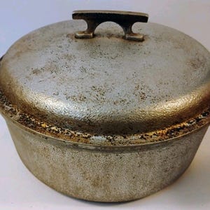 Puede incluir: Una olla redonda de metal color plata con tapa a juego. La olla cuenta con dos asas laterales y un asa central en la tapa. La superficie exterior tiene un aspecto texturizado y desgastado con signos visibles de uso y decoloración.