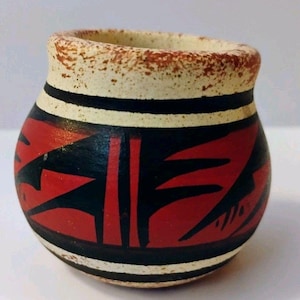 Peut inclure: Un petit pot en céramique arrondi présentant une base de couleur crème avec une texture mouchetée. L'extérieur est orné de motifs géométriques noirs audacieux et d'accents rouge vif. Une épaisse bande rouge mouchetée entoure le bord supérieur.