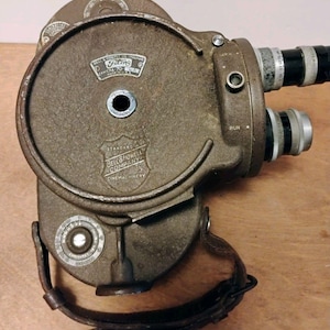 Pode incluir: Uma câmara de filmar vintage castanha Bell & Howell Filmo 70 com duas lentes, uma correia de couro e mostradores de exposição circulares. O corpo de metal apresenta texto gravado, incluindo Bell & Howell Company, Chicago e Camera 70 Made in U.S.A.