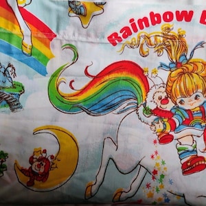 May include: A white fabric featuring a colorful Rainbow Brite print with the character riding a white horse with a rainbow mane. The design includes a yellow crescent moon, a small red house, a bridge, and the text Rainbow Brite in red letters.