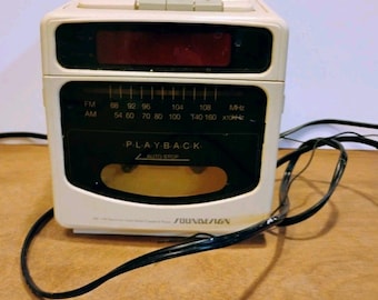 May include: A white Soundesign AM/FM clock radio and cassette player with a black front panel. The device features a digital clock display, a manual radio tuning dial with frequency markings, a cassette deck labeled PLAYBACK AUTO STOP, and a black power cord.