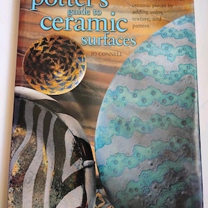 Puede incluir: Un libro titulado The Potter's Guide to Ceramic Surfaces de Jo Connell. La portada presenta un patrón circular marrón y dorado, una pieza de cerámica con rayas negras y blancas, y una gran forma ovalada texturizada azul y verde sobre fondo marrón.