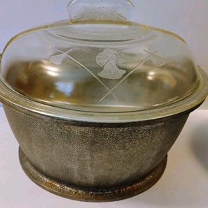 May include: A round metal cooking pot with a textured, dark bronze-colored exterior and two side handles. It features a clear glass lid with a central handle, decorated with an etched design of a knight's helmet positioned between two crossed spears.