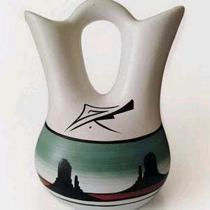 May include: A cream-coloured ceramic vase with a unique handle and a painted landscape design. The vase features a black geometric design and a green and black landscape scene with red and white stripes. The vase is approximately 20 cm tall.