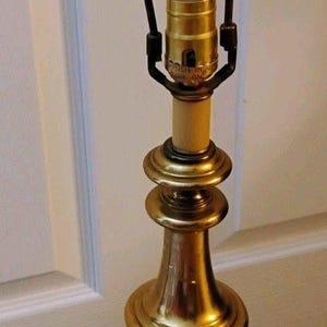 Puede incluir: Una base de lámpara de mesa de latón pulido con un diseño de pedestal redondeado y escalonado. La lámpara incluye un casquillo central con interruptor de botón y una lira metálica arqueada en la parte superior para la pantalla.