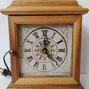 May include: A wooden mantel clock with a square frame and a brass handle on top. The clock face features black Roman numerals, ornate gold filigree patterns, and black hands. The text Old Master is printed at the bottom of the white clock face.