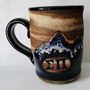 May include: A handcrafted ceramic mug with a black handle and rim. The mug features a mountain design with blue and white accents over a dark blue base. The mug has a brown and tan textured exterior and an oval emblem with Greek letters.