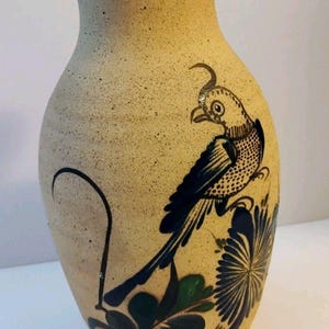 May include: A beige ceramic vase with a speckled texture, featuring a dark brown rim and a hand-painted design of a bird perched on a large flower. The bird and floral motifs are rendered in dark blue and green tones, with a dark band at the base.