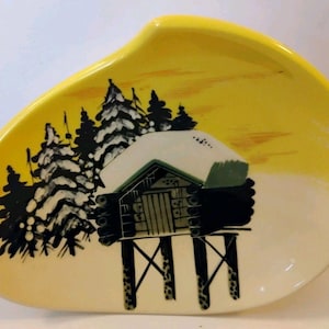May include: An asymmetrical ceramic dish with a bright yellow rim fading to cream. The center features a black and white illustration of a log cabin on stilts surrounded by snow-covered pine trees against a yellow sky background.