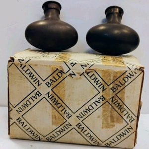 May include: Two dark brown, oval-shaped metal door knobs positioned above a rectangular cardboard box. The box is off-white with a repeating pattern of the word BALDWIN printed in black text inside diamond-shaped outlines across the surface.