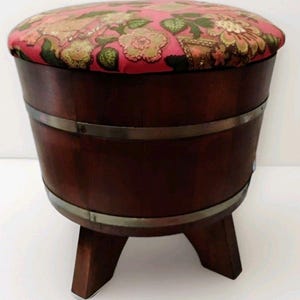 May include: A round wooden stool with a dark brown finish and two horizontal metal bands. The seat is upholstered in a pink fabric featuring a multi-colored floral pattern. The stool is supported by three short, sturdy wooden legs.
