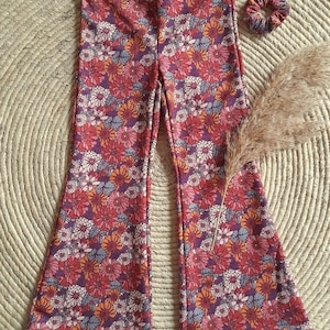 Könnte beinhalten: Eine Schlaghose und ein passendes Scrunchie, beide mit einem lebendigen Blumenmuster in Rot-, Rosa-, Orange- und Lilatönen. Die Hose ist aus weichem Stoff und hat einen elastischen Bund. Das Scrunchie ist aus dem gleichen Material.