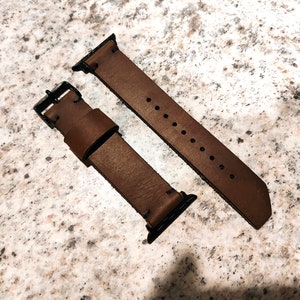 Puede incluir: Dos correas de reloj de cuero marrón con costuras negras y cierres metálicos. Las correas están diseñadas para Apple Watches.